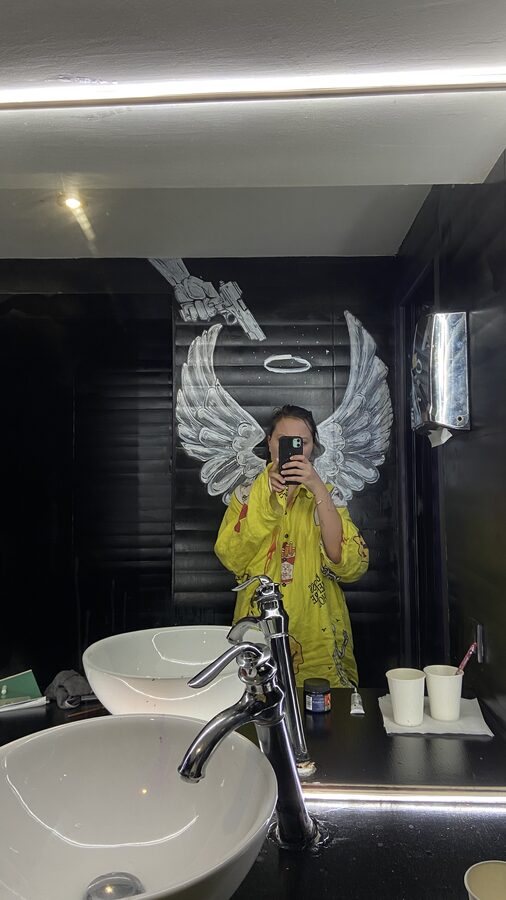 Mirror selfie in front of a painted wall mural of white wings and a halo